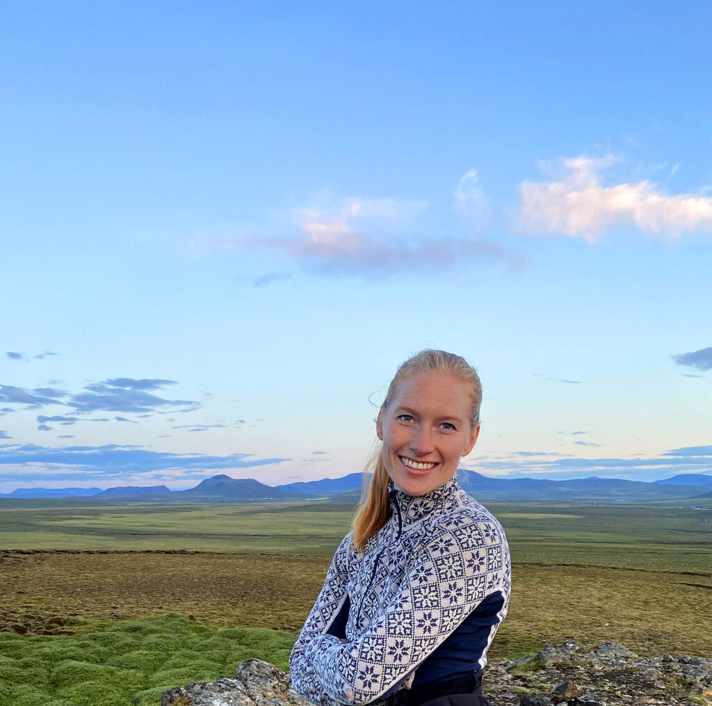 Smiling young woman, half figure, Icelandic views with grass, flat landscape, distant mountains and blue sky.