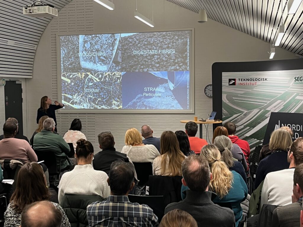 Conference room in white, a bit dark-ish, female and male audience watching a photo on a wall, listening to female presenter.